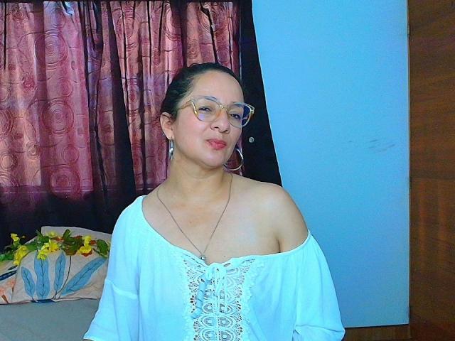 VictoriaRousse, with glasses and hoop earrings, wearing a white off-shoulder top, showing her bare shoulders and a delicate necklace, pouting slightly. Her tits are small and cute.