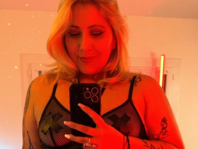 Blonde BlackWidowsI with wavy hair, red-tinted face, wearing black mesh lingerie with X-shaped pasties over tits, holding phone, tattoo on right arm, slight smile.