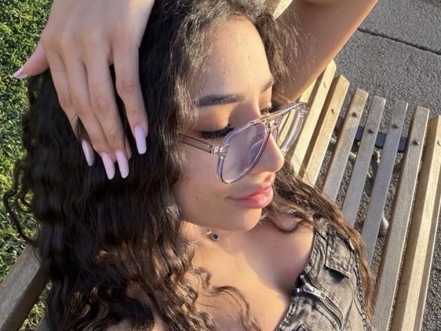 RamiraRowa, a young woman with long, wavy black hair, sits on a wooden bench in sunlight. She wears clear, cat-eye sunglasses with a small star on the right temple and has long, pink manicured nails. Her left hand rests on her head, and her right arm is relaxed. She wears a black, patterned top with a small, dark circular necklace. The green grass and pavement are in the background. Her eyes are closed, and her lips are slightly parted. The sunlight highlights her dark eyebrows and soft, tan skin.