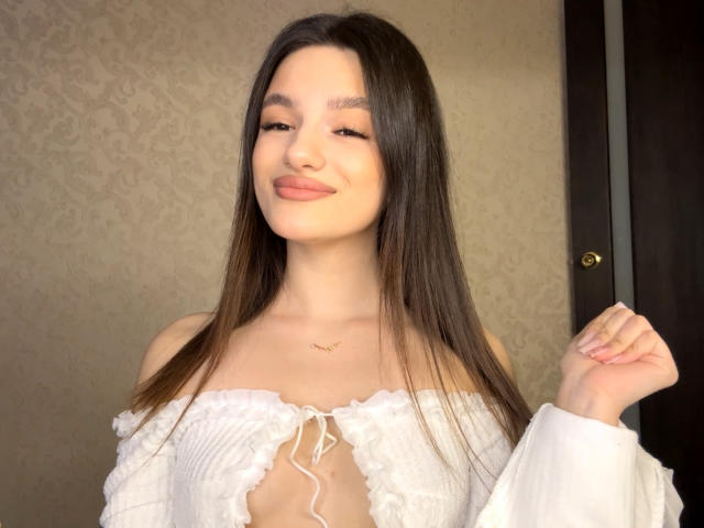 JinnyWalthers has long brown hair, a cute smile, and is wearing a white off-shoulder top with a lace-up front, showing a bit of her tits. She's holding her hair.