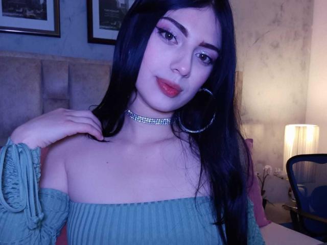 Isabella Rossi tiene el cabello largo y negro, cejas pobladas, y usa delineador de ojos llamativo con lápiz labial rosa. Lleva una blusa turquesa con un hombro descubierto, mangas abullonadas, un collar brillante y grandes aros. Su mano derecha toca su clavícula. La imagen tiene una luz azulada y fresca, que resalta su piel clara. La composición se centra en la parte superior de su cuerpo y su rostro. La textura de su blusa contrasta con la suavidad de su piel y su cabello. Su expresión es ligeramente curiosa, con una sonrisa sutil..