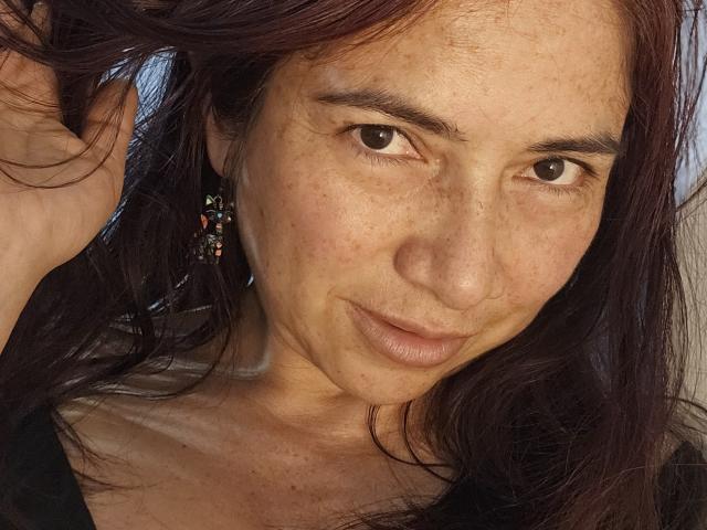 AlessiaRed has freckled, slightly sunburned skin with dark brown eyes, wavy red-brown hair, and a subtle smile. She's wearing colorful earrings, looking directly at the camera.