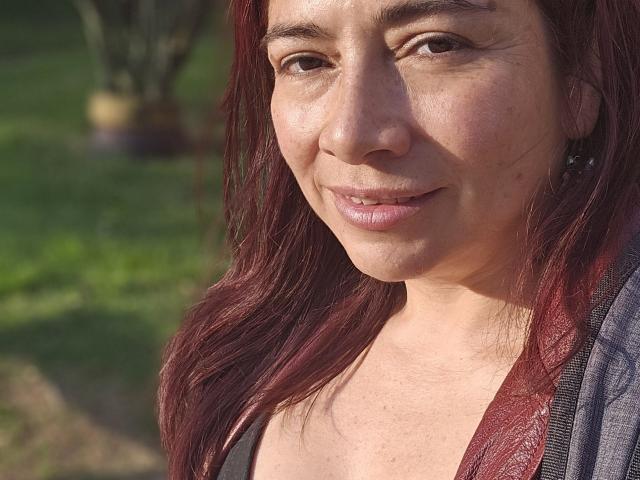 AlessiaRed with red hair, fair skin, and small tits, smiling softly, wearing a gray jacket, in a sunlit outdoor setting. She has subtle makeup and earrings.