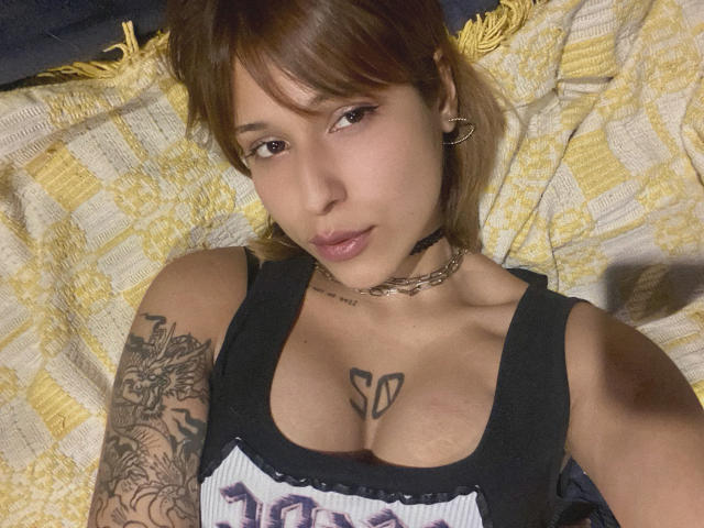 KiaraHernandezX lies on a patterned yellow and white blanket. She has light brown skin, straight brown hair with bangs, and wears small hoop earrings. Her brown eyes are slightly open, and she has a small, pinkish lip. She wears a black tank top with "100%" in purple letters, and a chain choker. A tattoo of the number "50" is on her chest, and a detailed black and gray tattoo covers her right shoulder. The image is a close-up selfie.