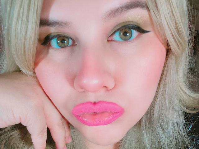 Akkanee's face with blue eyes, blonde hair, pink lipstick, and black eyeliner. She's resting her chin on her hand, showing a nose ring. Her skin is fair.