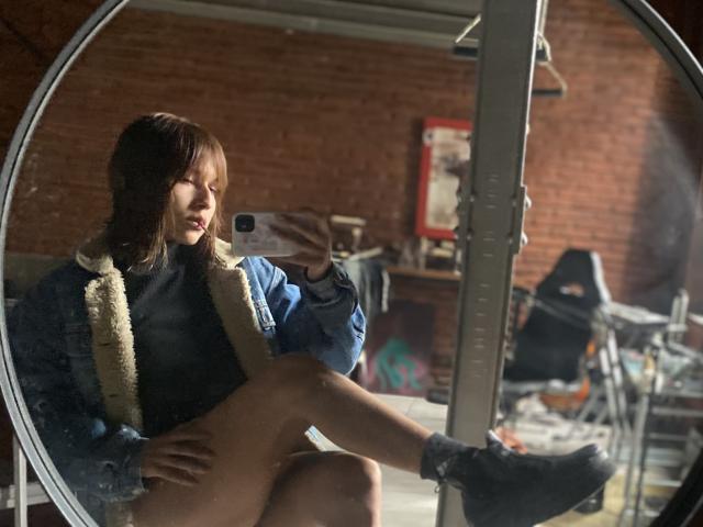 KiaraHernandezX sits in a circular mirror, taking a selfie. She wears a blue jacket with a fluffy collar, a black shirt, and black socks. Her brown hair is loose, and she has a focused expression. Her right leg is bent, foot resting on the mirror's edge. The room has a brick wall, scattered furniture, and a red-framed poster in the background. Natural light highlights her face and jacket. Reflection and shadow play add depth to the image.