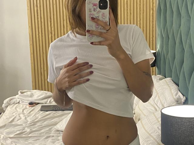 KiaraHernandezX takes a mirror selfie in a bedroom. She's wearing a white, short-sleeved ribbed crop top, lifting it to reveal her flat, tan midriff. Her right hand holds a phone with a pink floral case, partially covering her face. Her left hand rests on her chest. She has a tattoo on her left forearm. The bed has white, textured pillows and a blue, tufted headboard. A blue lamp is on the right. Her nails are painted dark purple.