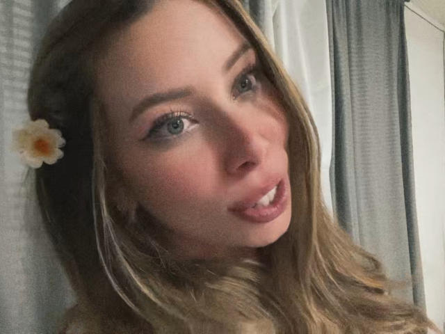 JennyLawry's face with long blonde hair, a white flower in her hair, and a slightly open mouth showing a small bit of her tongue. She has blue eyes and pink lips.