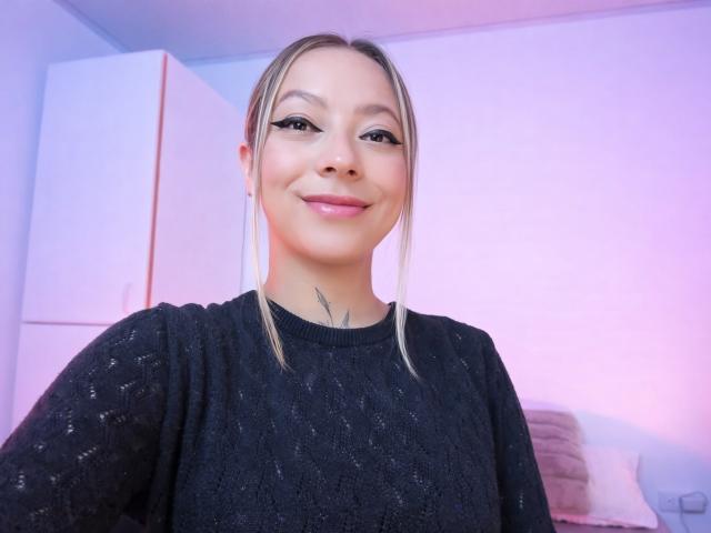 KayleeM has a light skin tone, straight blonde hair in a simple side part, and wears a black textured sweater. She has a small tattoo under her chin and is smiling softly with pink lipstick. Her eyeliner is winged, and she has subtle, natural-looking makeup. The background has a pinkish-purple light, a white door, and a pillow on a bed. The image is a close-up selfie.