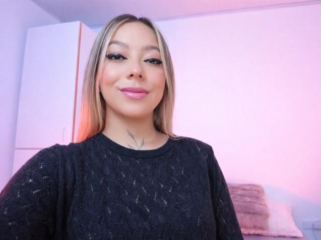 KayleeM with blonde hair, pink lipstick, and black sparkly sweater, smiling at the camera, showing a small tattoo on her neck. Her tits are not visible.