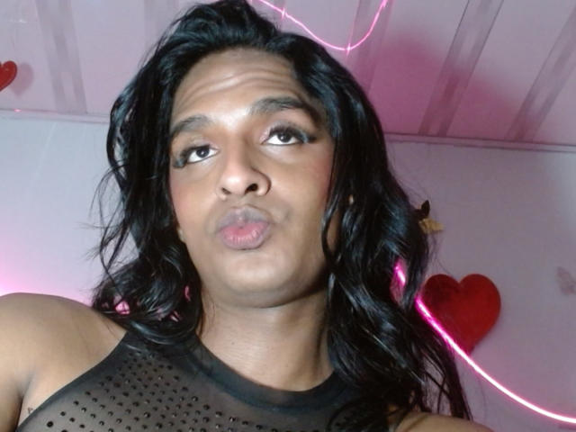 Close-up photo of IdiiaLatina with medium brown skin, dark wavy hair, and brown eyes. She's making a pouty face with slightly pursed lips. She wears a black, sheer, polka-dotted sleeveless top. Pink neon lights are visible in the background, and a red heart decoration is on the right. Her hair is loose and falls around her face. The lighting is even, highlighting her facial features. The ceiling is white with visible panels.
