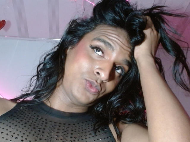 Close-up photo of IdiiaLatina with long, wavy black hair, holding a section of it with her right hand. She has dark brown skin, thick eyebrows, and is puckering her lips slightly. She's wearing a sheer black top with small polka dots. Her eyes are looking to the right, and she has a slight frown. The lighting is soft, with a pinkish hue in the background. Her makeup includes bold eyeliner. The image has a casual, selfie-like quality.