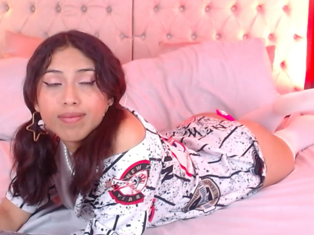 MyaaTyler is lying on a bed with white sheets. She has long, wavy, dark brown hair and is wearing a white, off-shoulder, graphic print dress with black and red splashes. She's also wearing white knee-high socks. Her eyes are closed, and she has a slight smile. Star-shaped earrings are in her left ear. The bed's tufted, white headboard is in the background. The lighting is pink. She has a medium skin tone.