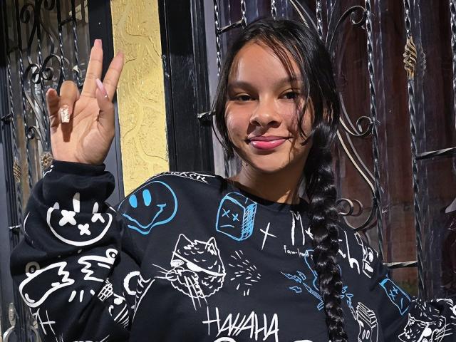 Yurimy with dark hair in a braid, wearing a black shirt with blue and white graffiti-style designs, making a peace sign with her right hand, pink lipstick.