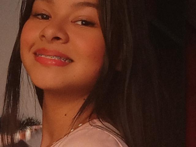 Yurimy with straight black hair, brown eyes, and shiny pink lips, smiling slightly with metal braces showing. She has a small mole on her neck.