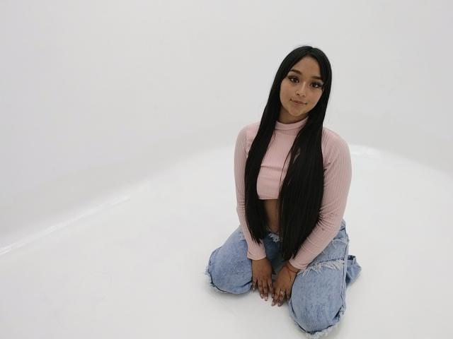 EilynTatiana with long black hair, pink ribbed crop top, and blue jeans, kneeling on white floor, hands on knees, smiling softly, showing small, cute ass and medium tits.