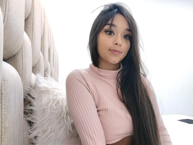 EilynTatiana with long black hair, pink ribbed crop top, sitting on beige tufted sofa, white fluffy pillow on left, smiling softly, light skin, brown eyes.