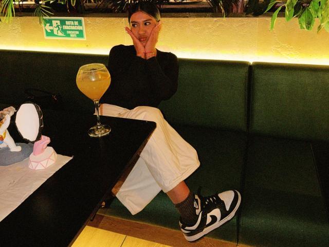 ThaliaAres with hands on face, black long-sleeve shirt, white pants, black and white sneakers, sitting on green booth, glass of yellow drink, black table, cute pink toy.