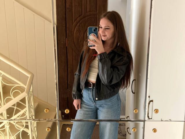 PamelaXu with long brown hair, black leather jacket, white top, and blue high-waisted jeans, taking a mirror selfie, showing a subtle smile, and holding a blue phone.