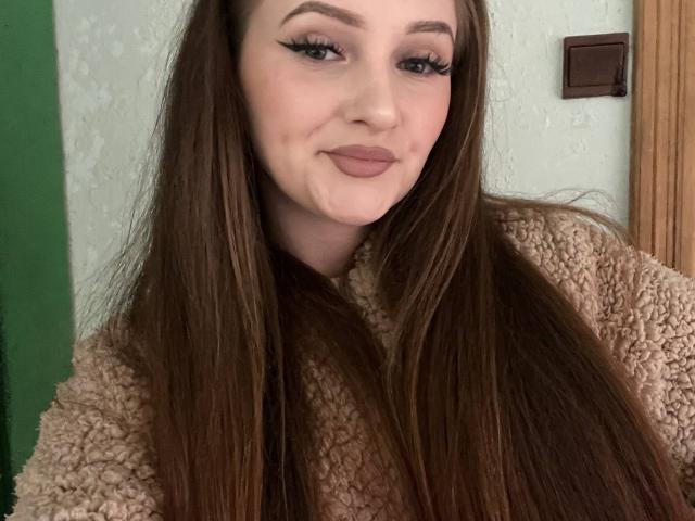 PamelaXu with long brown hair, wearing a fluffy beige sweater, has subtle makeup with winged eyeliner and pink lipstick, smiling slightly. Her tits are not visible.