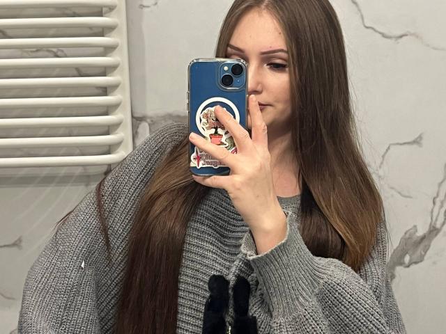PamelaXu with long brown hair, gray sweater, and blue phone case with a white and red graphic, taking a mirror selfie, showing her nose and partial face.