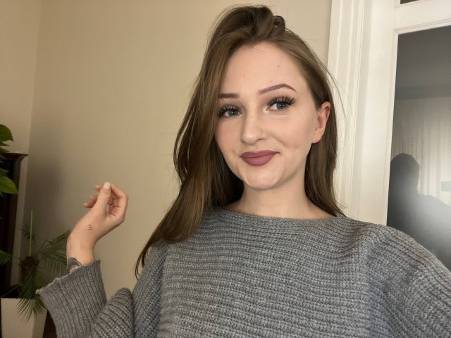 PamelaXu with long brown hair, wearing a grey ribbed sweater, has a cute smile, full eyelashes, and pink lipstick, holding her hair up. Her tits are medium-sized.