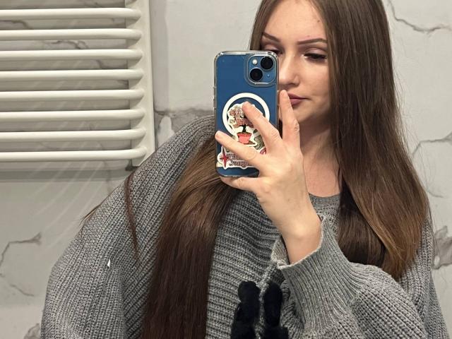 PamelaXu with long brown hair, wearing a gray knitted sweater, taking a mirror selfie with a blue phone featuring a colorful pop-up button. Her face shows a slight smile.