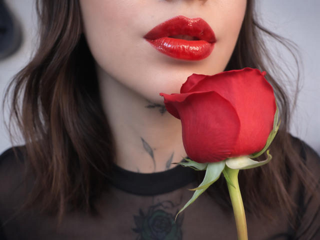 Close-up of KayleeM's face, focusing on her glossy red lips and the red rose she holds near her mouth. KayleeM has fair skin and dark brown, wavy hair. She wears a sheer black top, revealing a tattoo on her chest. The rose's vibrant red petals contrast with the green stem. The image emphasizes the rose's texture and KayleeM's shiny lipstick. Slight shadow on her chin.