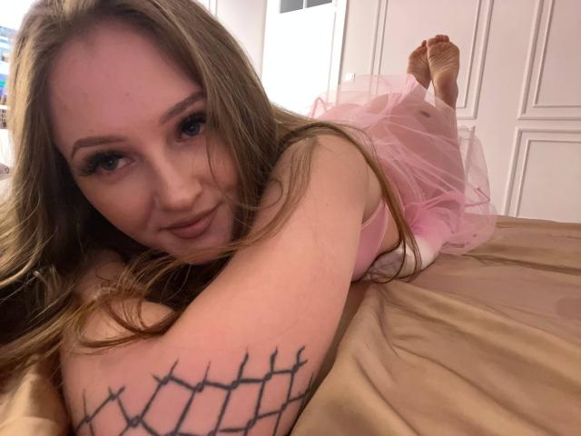 Photograph of PamelaXu lying on a bed. She has long, wavy blonde hair, light skin, and is wearing a pink, sheer, sleeveless dress with a tutu-like bottom. Her eyes are slightly squinted with a subtle smile. She has a black, mesh-like tattoo on her right forearm. Her feet are up in the air, and she's on a beige bed. The image is focused on her upper body and face, with soft lighting. The composition is casual and intimate. Her makeup is subtle, with long, dark eyelashes. The background is mostly out of focus.