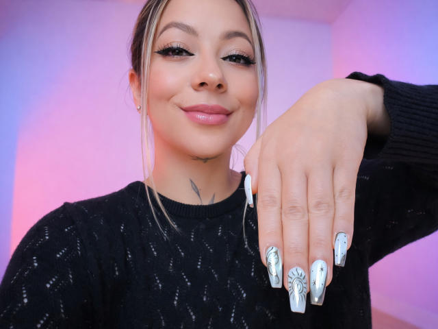 KayleeM with long blonde hair, black sweater, and white nails with green leaf designs, smiling, hand resting on her chin, subtle nose ring, and a small tattoo under her chin.