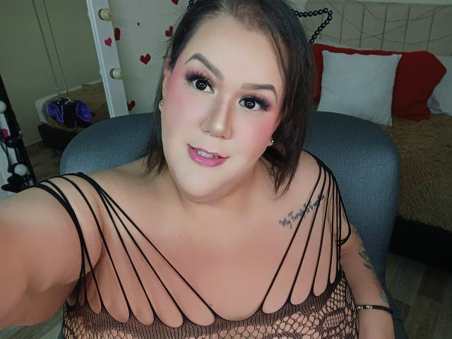 GiiannaMorrys con il trucco rosa, orecchie da gatto nere e una maglia a rete, che mostra il suo seno prosperoso e il tatuaggio sul braccio destro, seduta su una sedia..