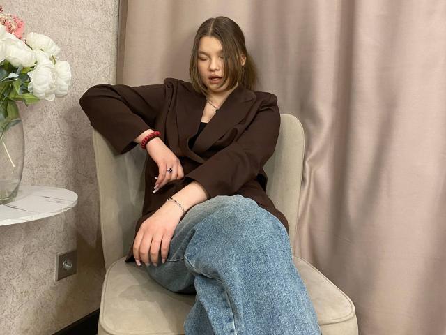 IvyCeovantes with brown jacket, black top, and blue jeans, sitting on beige chair, eyes closed, right arm resting on knee, left hand on chair, wrist bracelet, and flower vase.