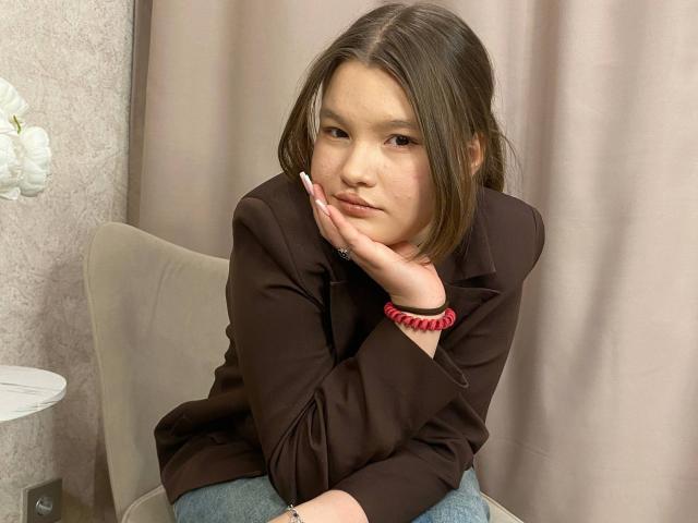 IvyCeovantes, a young Asian woman with straight brown hair, sits on a beige chair, resting her head on her left hand. She wears a brown blouse and blue jeans, with a red beaded bracelet on her right wrist. Her right hand is slightly raised, showing white manicured nails. She looks directly at the camera with a neutral expression. The background has a beige curtain. The image is clear and focused.