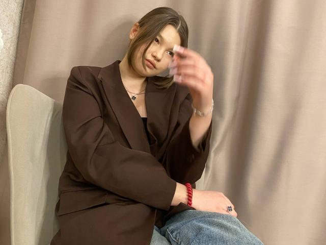 IvyCeovantes, a young woman with fair skin and brown hair, sits on a beige chair. She wears a brown blazer over a black top and blue jeans. Her right hand is raised, slightly blurred, suggesting movement. She has a neutral expression and a small necklace with a pendant. Her left wrist has a red bracelet. The background is a plain, beige curtain. The composition is simple and focused on IvyCeovantes. Her hair is styled in a side part. The image has a casual, everyday style.