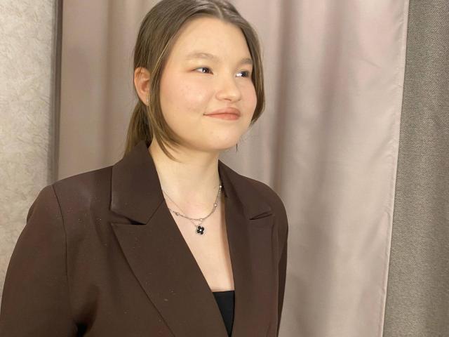 IvyCeovantes with light skin, brown hair in a ponytail, brown blazer, black top, small black necklace with a pendant, slight smile, looking to the side.