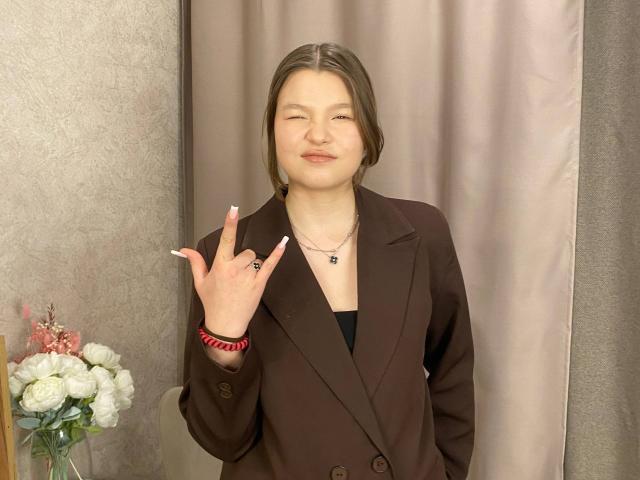 IvyCeovantes with short brown hair, wearing a brown blazer, black top, holding up a finger with a ring, and a necklace, standing in a room with a flower vase.