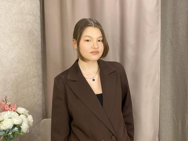 IvyCeovantes, an Asian woman with fair skin and straight brown hair in a side part, stands in a neutral, indoor setting. She wears a brown blazer over a black top, with a small black pendant around her neck. Her expression is calm, with a slight, neutral smile. A bouquet of white and pink flowers is on a table to her left. The background features a grey curtain and a textured wall. The composition is centered, highlighting IvyCeovantes' upper body and face. The lighting is even, with no harsh shadows.
