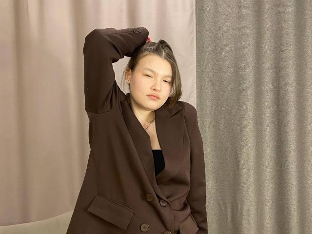 IvyCeovantes stands with her right arm raised, hand resting on her head. She wears a brown, buttoned blazer over a black top. Her eyes are half-closed, giving a relaxed, slightly bored expression. She has fair skin, light brown hair, and small earrings. The image is well-lit, with a subtle shadow on her face. The background features two textures: a pink curtain on the left and a grey curtain on the right. IvyCeovantes is centered in the frame.