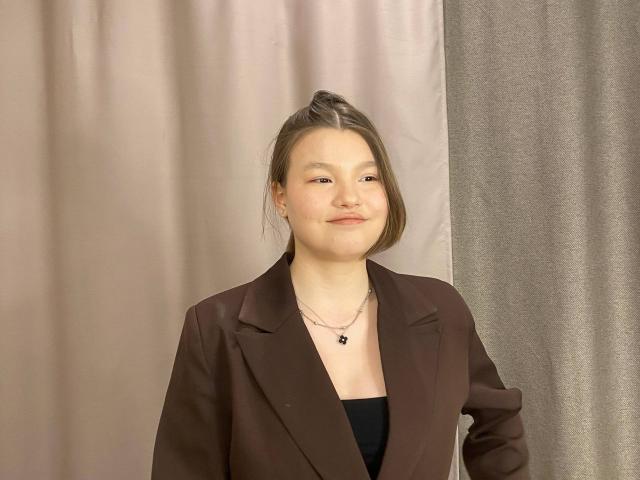 IvyCeovantes, a young woman with fair skin and a subtle smile, stands against a simple backdrop. She has a shoulder-length brown bob and wears a brown blazer over a black top. A thin necklace with a small black pendant hangs around her neck. The blazer's fabric is smooth, and her hair is slightly messy. The overall composition is straightforward, with a neutral color palette that emphasizes IvyCeovantes' calm and confident demeanor. The image has a casual, realistic style.