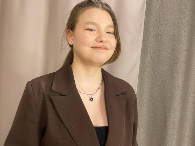 IvyCeovantes, a young woman with light skin and brown hair in a ponytail, stands with a subtle, confident smile. She wears a dark brown blazer over a black top, with a simple silver necklace featuring a small black pendant. Her eyes are slightly squinted, and her face has a natural, relaxed expression. The composition is centered, with soft, muted background colors. The overall style is casual and approachable.