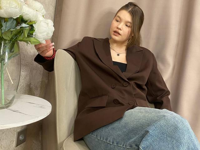 IvyCeovantes sits in a beige armchair, wearing a brown double-breasted jacket and blue jeans. She holds a pink bracelet in her right hand, with white roses in a glass vase on a white round table to her left. IvyCeovantes has light brown hair, a fair skin tone, and a relaxed expression. The background features beige curtains. The composition focuses on IvyCeovantes' casual, comfortable pose and the contrast between the brown jacket and blue jeans. The image uses natural light, highlighting IvyCeovantes' relaxed demeanor.