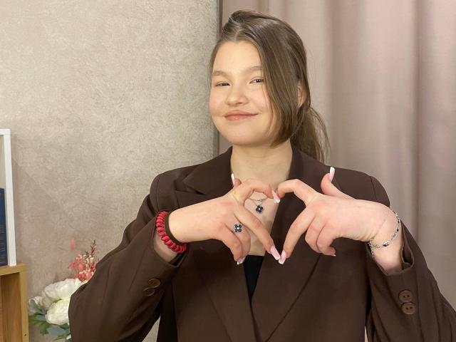 IvyCeovantes stands in a room with beige walls and pink curtains. She has light skin and brown hair in a loose ponytail. She wears a brown blazer and has pink nail polish. IvyCeovantes forms a heart shape with her fingers, showing a black ring on her right index finger and a silver ring on her left ring finger. She also wears a red bracelet on her left wrist. The image is a photograph.
