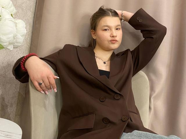 IvyCeovantes, a young woman with light skin and brown hair in a simple, pulled-back style, sits in a beige chair. She wears a brown double-breasted coat with large buttons and a black top underneath. Her right arm is raised, resting on the chair's back, while her left arm rests on the chair's armrest. She has a red scrunchie on her left wrist and a black ring on her left hand. Her nails are long and painted white. A silver necklace with a small pendant is visible. Her expression is neutral, slightly pouting. The background is a beige curtain. White flowers are partially visible in the top left corner.