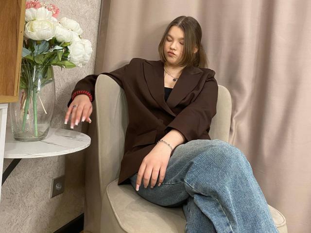 IvyCeovantes sits in a beige armchair, wearing a brown jacket over a black top and blue jeans. Her eyes are closed, and her right arm rests on the chair's armrest, with her hand slightly raised. She has light brown hair, and a red bracelet is visible on her wrist. Her left arm is crossed over her lap. A small round white table on the left holds a clear vase with white roses. The background features a beige curtain. The composition is balanced, with IvyCeovantes' relaxed posture and casual clothing creating a calm, everyday scene. The textures of the armchair and jacket contrast with her smooth skin and denim.
