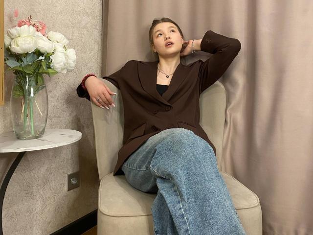 IvyCeovantes is sitting in a beige chair, leaning back with her right arm resting on the chair's back and her left arm on the chair's armrest. She's wearing a brown blazer, black top, and blue jeans. She has light brown hair pulled back, red lipstick, and a red bracelet on her left wrist. A white vase with white and pink flowers is on a small round table to her left. The background has beige curtains and textured wallpaper.