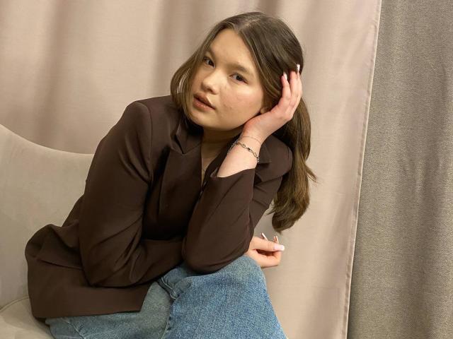 IvyCeovantes with brown hair, brown blouse, and blue jeans, sitting on a chair, hand on head, looking slightly bored, her ass subtly pressed against the chair.