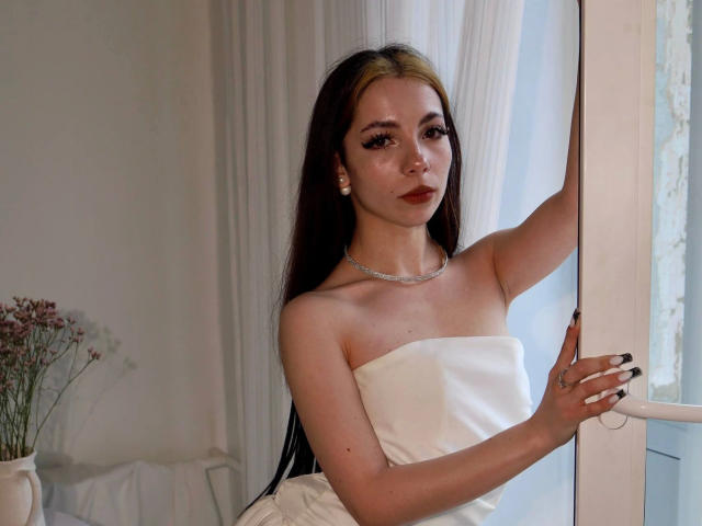 LiaPeach-hot with long dark hair, white strapless dress, silver necklace, and pearl earrings, standing by a window, touching it with her right hand, looking at the camera.