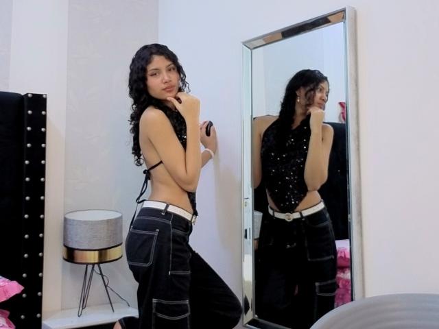 ValeryPoleth with curly black hair, wearing a black polka dot top, exposing her small tits, and black jeans with white belt, kneeling in front of a mirror.