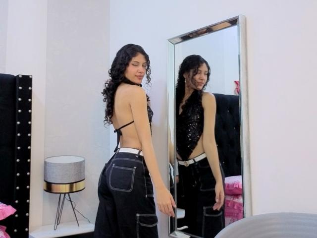 ValeryPoleth, a young woman with long curly black hair, stands in a room, facing a mirror. She wears a black sequined halter top and black denim pants with white straps. Her back is to the camera, but she looks over her shoulder at the mirror, showing her reflection. The mirror has a light frame, and a black, tufted headboard is partially visible. A small, modern lamp is on a white shelf to the left. The room has white walls and a pink pillow on a bed. The overall composition highlights ValeryPoleth's reflection and the contrast between her black top and denim pants.