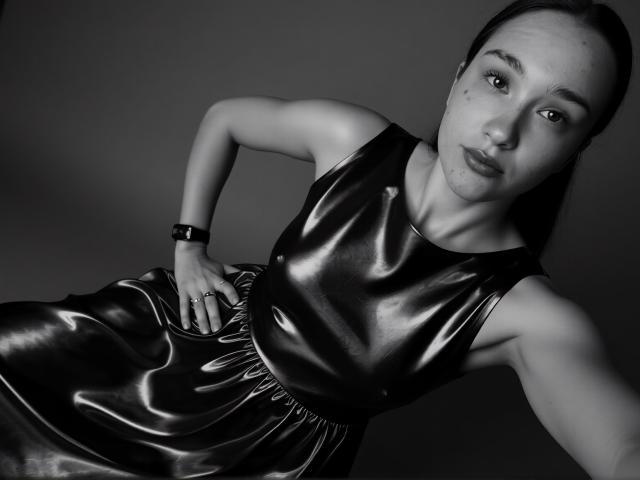 VertieTllo in a shiny black latex dress, leaning forward with one hand on her hip, wearing a wristwatch, black hair pulled back, and a slight pout. Black-and-white selfie.